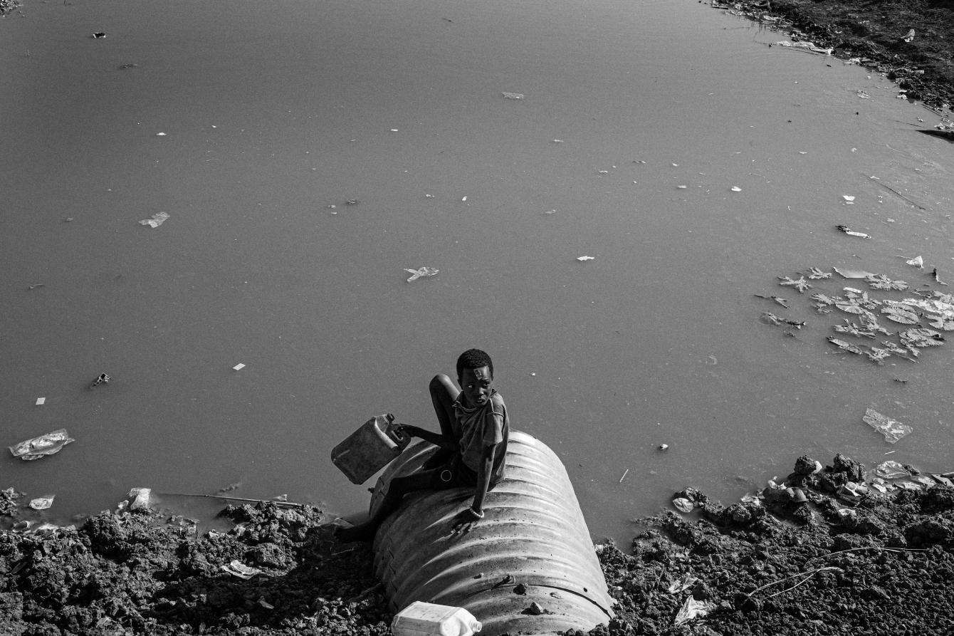 A boy sitting on a sewer pipe by a body of water.