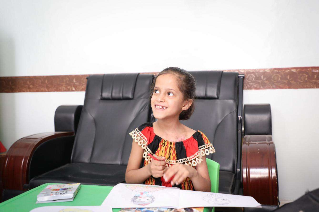 A seven year old girl in a bright dress, smiling, sitting at a table with a crayon in her hand.