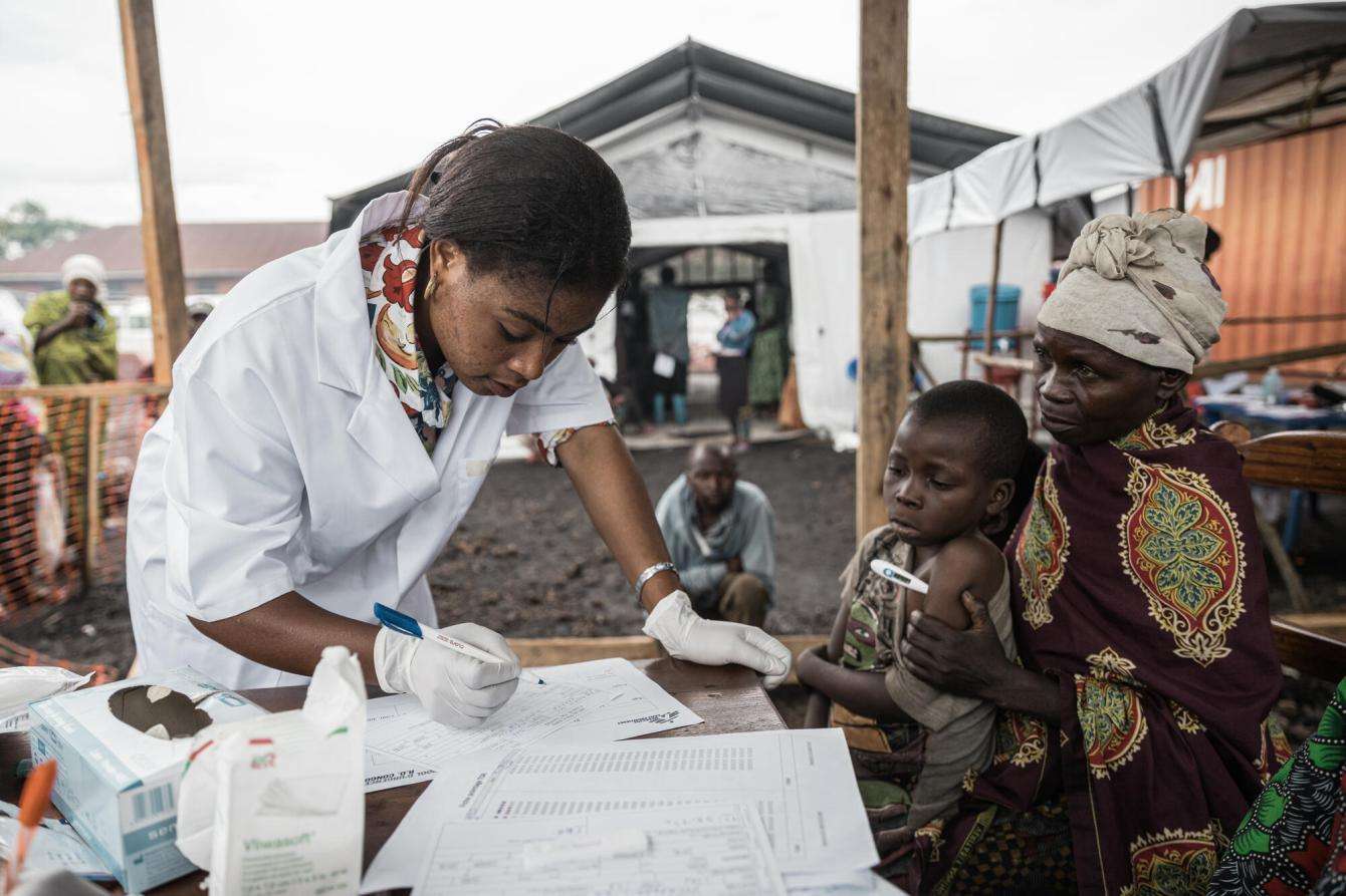 Woman in white lab coat fills paperwork for child and mother patients at MSF clinic in a displacement camp in Democratic Republic of Congo