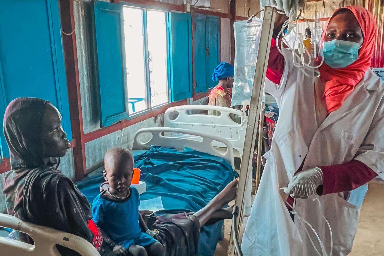 MSF medical staff member wearing white lab coat and headscarf treats a patient sitting in hospital bed in White Nile state, Sudan.