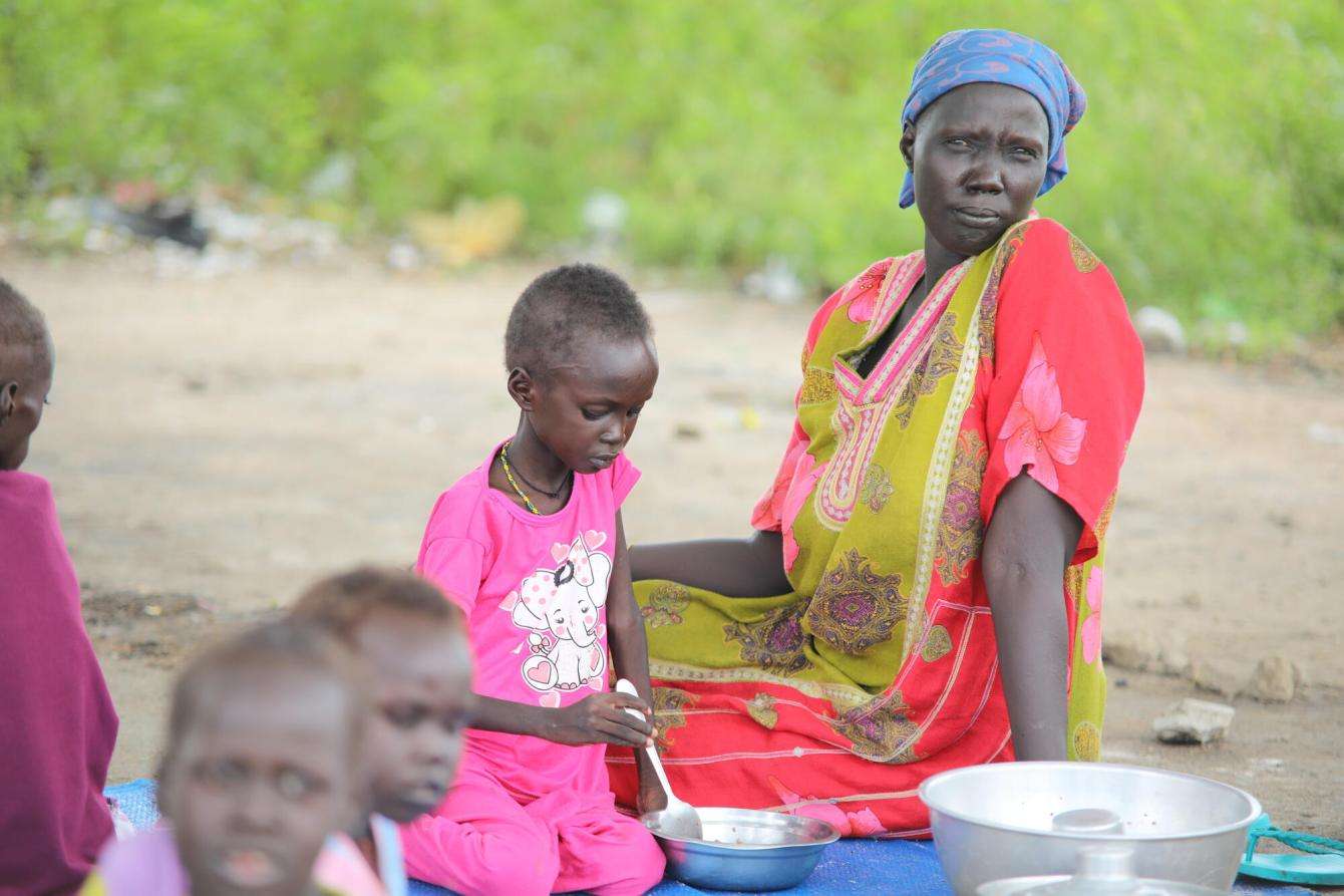 A mother sits outside with her children—MSF patients—as they prepare meals at thet Renk Civil Hospital
