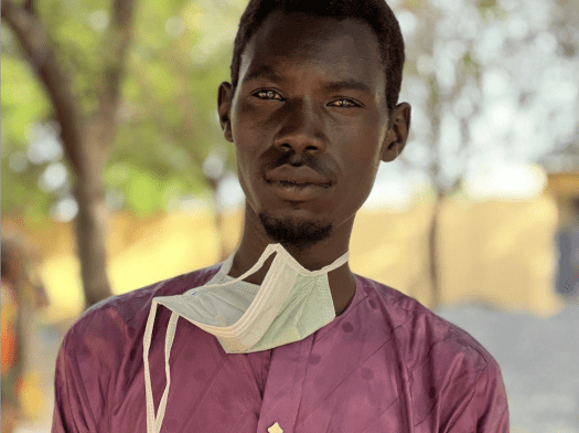 young man with mask tied around neck stands under trees in Chad after fleeing Sudan