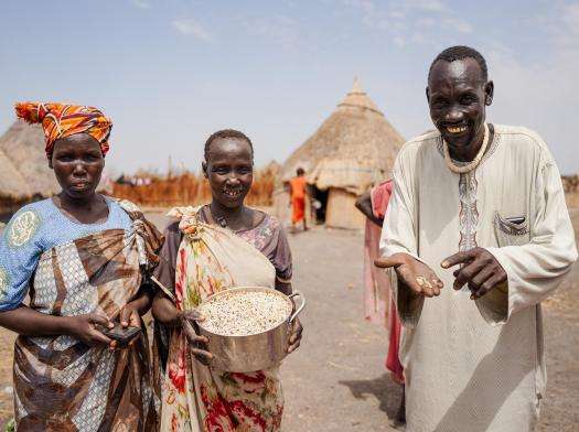 Three people stand with pots of rice in Aree, Akoka County, South Sudan, under blue sky in front of huts