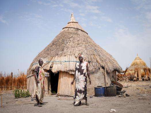 Women stand in front of a hut in Dentiuk village, South Sudan with blue sky